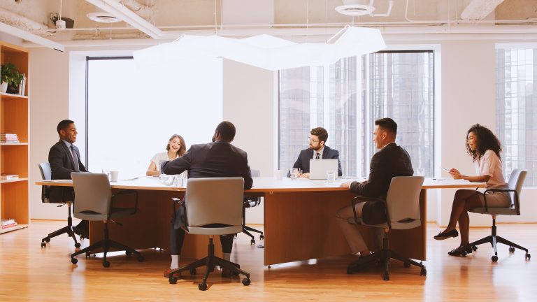 Six people in business attire sit around a large conference table in a modern office with big windows, engaged in an Executive Roundtable discussing market insights for May 2026.