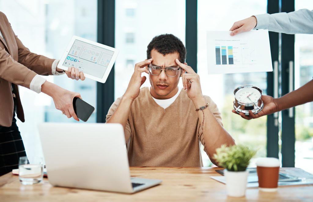A stressed man sits in the middle of a desk with a laptop, holding his temples. Multiple hands surround him with a tablet, phone, clock, and documents, illustrating overwhelm and multitasking at work.