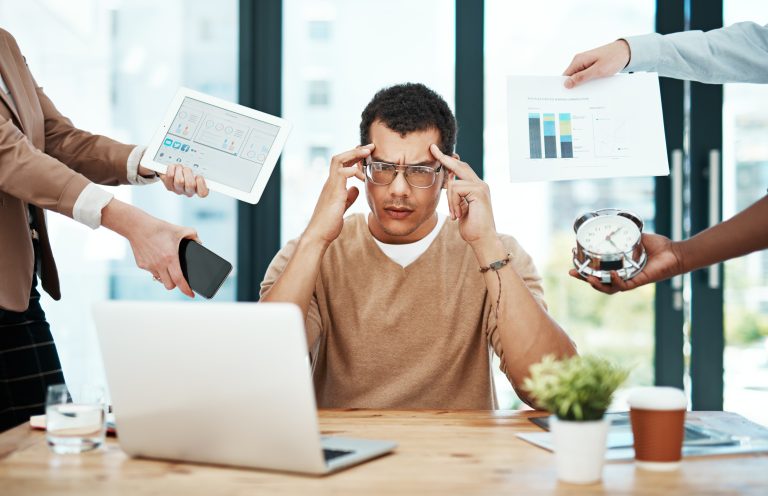 A stressed man sits in the middle of a desk with a laptop, holding his temples. Multiple hands surround him with a tablet, phone, clock, and documents, illustrating overwhelm and multitasking at work.