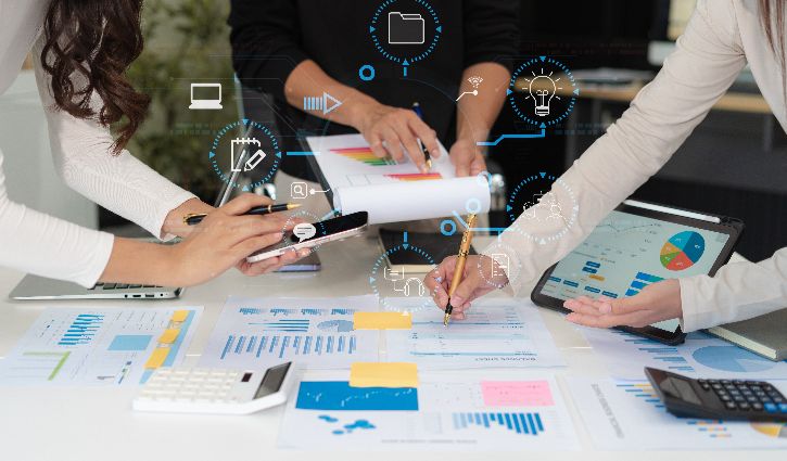 Three people analyzing business charts and graphs on paper, using a tablet, calculator, and pens at a white desk with digital business icons overlaid, representing consulting teamwork and data analysis.