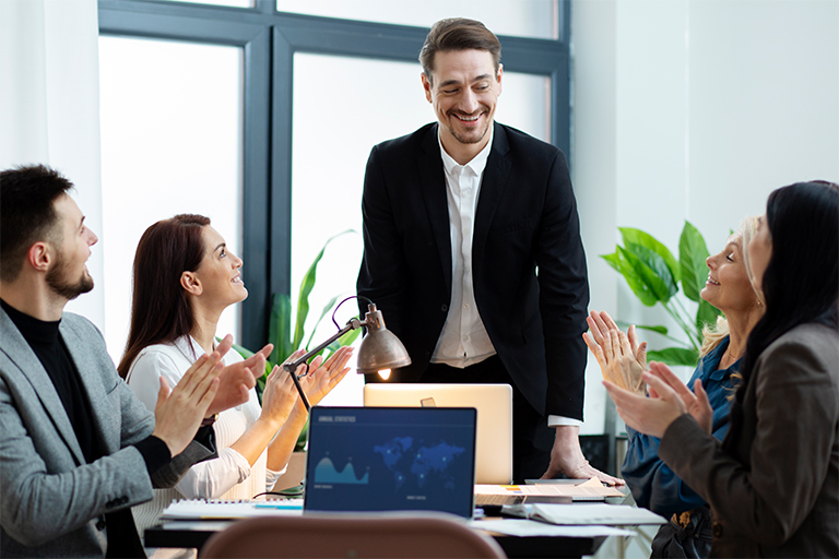 Four colleagues in business attire are gathered in an office, smiling and applauding a standing man in a suit, suggesting a successful meeting or celebration of leadership development. Laptops and plants are visible on the table, highlighting an environment focused on building teams.