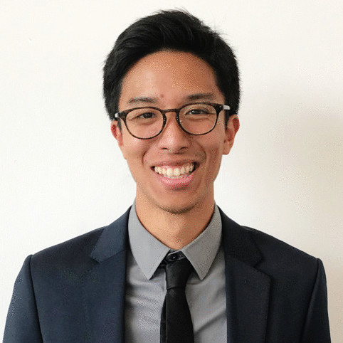 Mick Trinidad, a young man with short black hair and glasses, smiles at the camera. He is wearing a dark suit jacket, a gray dress shirt, and a black tie, standing against a plain white background.