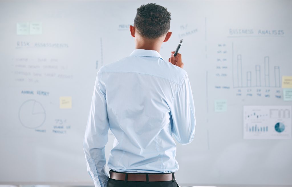 A man in a light blue shirt stands facing a whiteboard filled with charts, graphs, and notes, holding a marker and appearing to analyze or plan information related to Account Growth and Sales Performance.