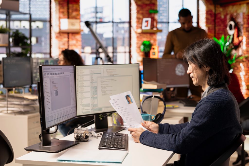 A person sits at a desk in a modern office, reviewing a printed resume while looking at a computer screen displaying tech hiring software. Other people work in the background, and large windows let in natural light.