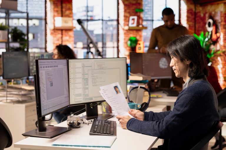 A person sits at a desk in a modern office, reviewing a printed resume while looking at a computer screen displaying tech hiring software. Other people work in the background, and large windows let in natural light.